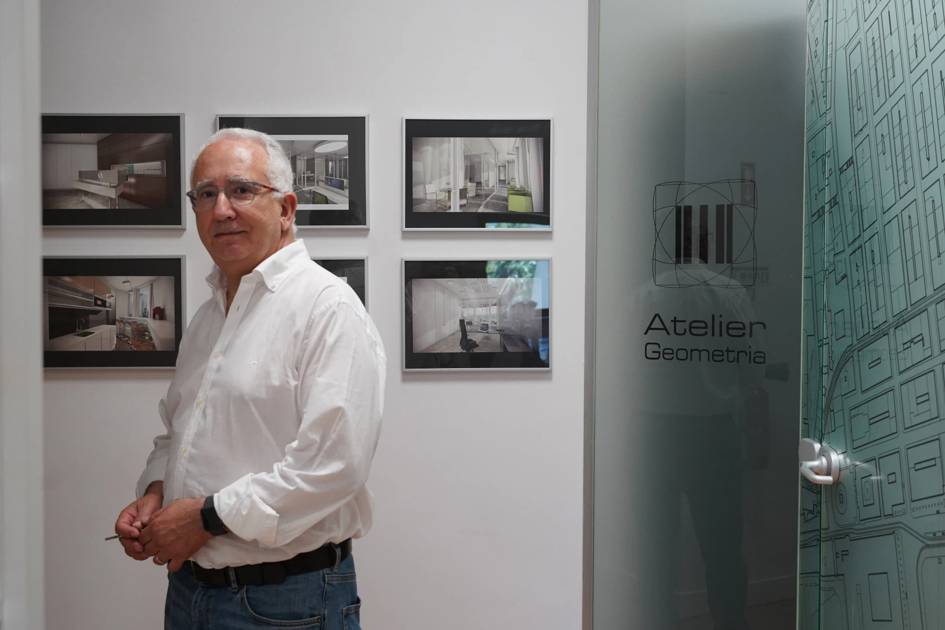 An architect in the studio, beside the wall of framed project images and the glass door etched with the Atelier Geometria mark